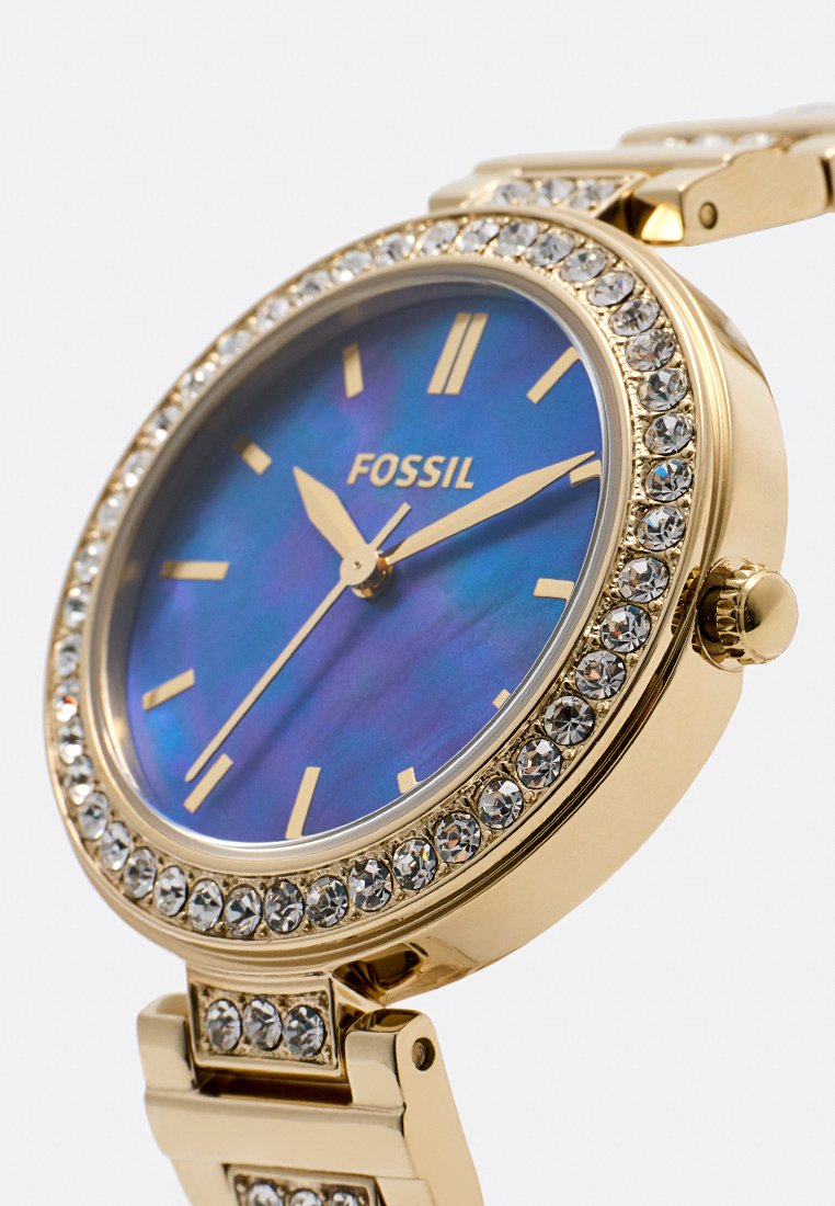 Montre d'or avec un cadran en nacre bleue, accents en cristal autour de la lunette, et index et aiguilles dorés. Design de bracelet en métal.