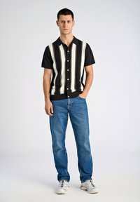 Manskjorta med kort ärm i svarta och krämvita vertikala ränder, med knäppning fram och platt krage, ihop med blå denimjeans och vita sneakers.