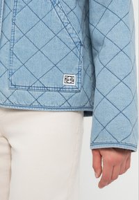 Ανοιχτό γαλάζιο τζην μπουφάν με ραφές σε αντίθετο σκούρο χρώμα, που διαθέτει μπροστινή τσέπη και μικρή ετικέτα με το εμπορικό σήμα.