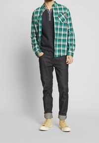 Grøn og blå ternet skjorte med knapper over en grå striktrøje, mørke jeans og lysegule sneakers. Afslappet pasform med op rullede ærmer.