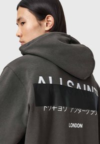 Kapuzensweatshirt aus dunkelgrauem Stoff mit weißem Text auf dem Rücken. Weiche Textur; lockere Passform, mit einem Akzent aus Schwarz, der einen Teil des Textes verdeckt.