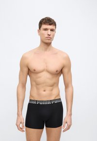 Νέος άνδρας με κοντά σγουρά μαλλιά, φοράει μαύρα εσώρουχα boxer Puma, στέκεται μπροστά σε απλό ανοιχτό γκρι φόντο.