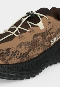 Καφέ και μαύρο παπούτσι trail running Salomon με μπεζ σύστημα γρήγορου δέσιμου και υφή στην σόλα, εμφανιζόμενο από πλάγια γωνία.