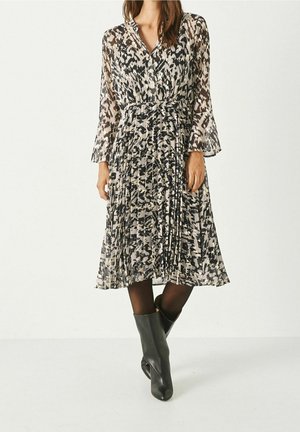 Femme portant une robe midi plissée à manches longues, noire et blanche à motifs, associée à des collants noirs et des bottines noires.