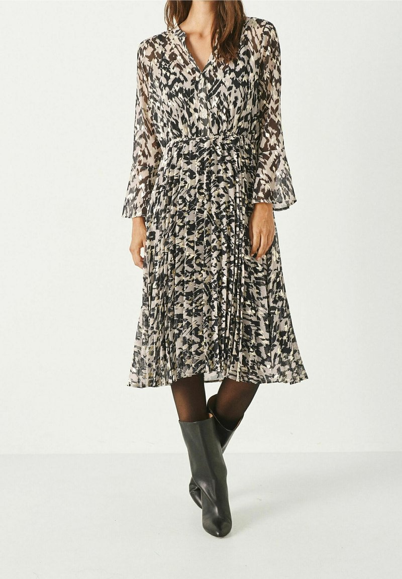 Femme portant une robe midi plissée à manches longues, noire et blanche à motifs, associée à des collants noirs et des bottines noires.