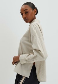 Light beige striped shirt with a relaxed fit, long sleeves, and collar. Features side slits and subtle multicolored stripes.