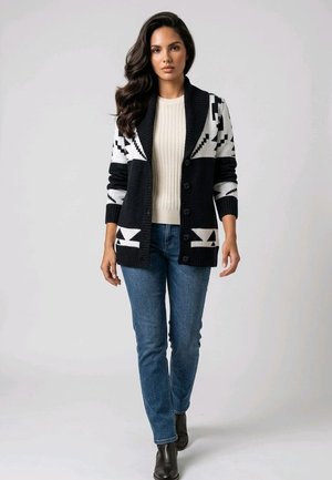 Mujer con cabello largo y oscuro lleva un cárdigan con patrón en blanco y negro, jersey crema de canalé, vaqueros azules y botas negras hasta el tobillo, de pie con expresión neutral.