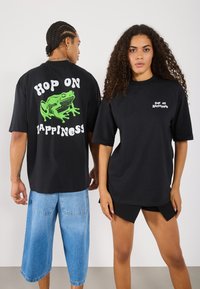 Melni oversize T-kreklus ir zaļš vardes grafiskais attēls un teksts "HOP ON HAPPINESS" aizmugurē, ar mazāku tekstu priekšpusē.