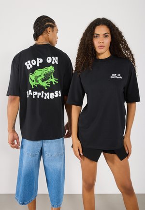 Schwarze übergroße T-Shirts mit einem grünen Frosch-Motiv und dem Text "HOP ON HAPPINESS" auf dem Rücken, sowie einem kleineren Text auf der Vorderseite.