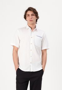 Camisa de manga corta, de color claro, hecha de tela suave que cuenta con un bolsillo en el pecho con un acento azul y un cierre de botones.