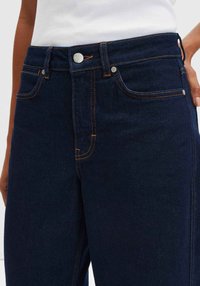 Jeans in denim blu scuro con vita alta, cuciture arancioni, design a cinque tasche, rivetti metallici e chiusura con bottone argentato.