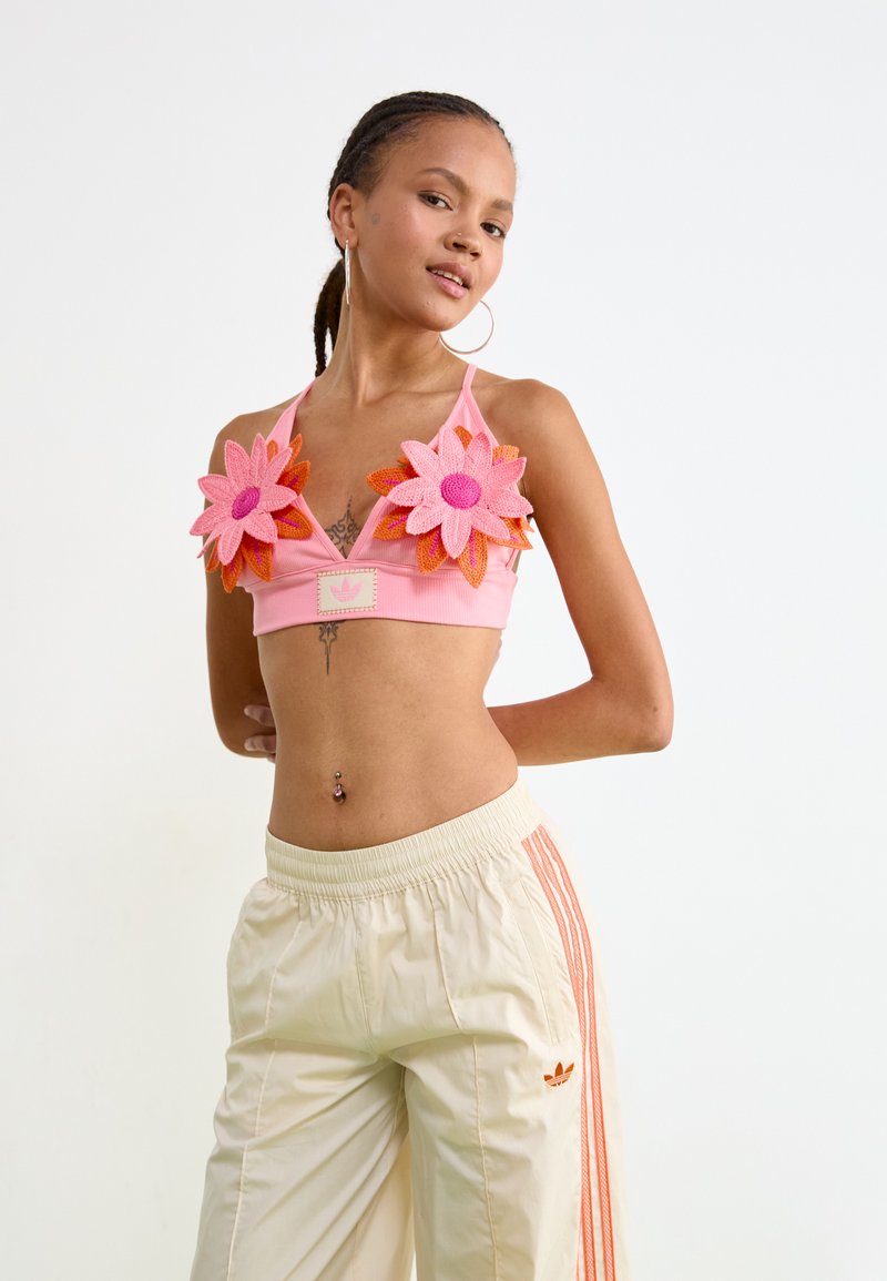 Różowy crop top z 3D kwiatowymi zdobieniami w odcieniach pomarańczowym i różowym. W zestawieniu z kremowymi dresami z pomarańczowymi paskami bocznymi i logo.