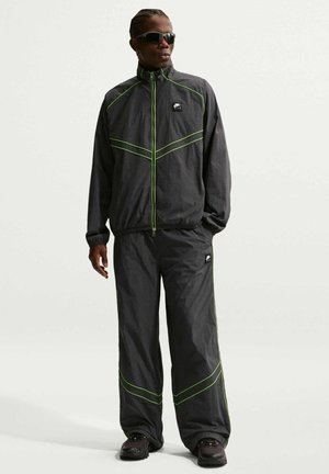 Uomo con tuta nera con rifiniture verde neon, scarpe da ginnastica nere e occhiali da sole scuri, in piedi davanti a sfondo bianco.