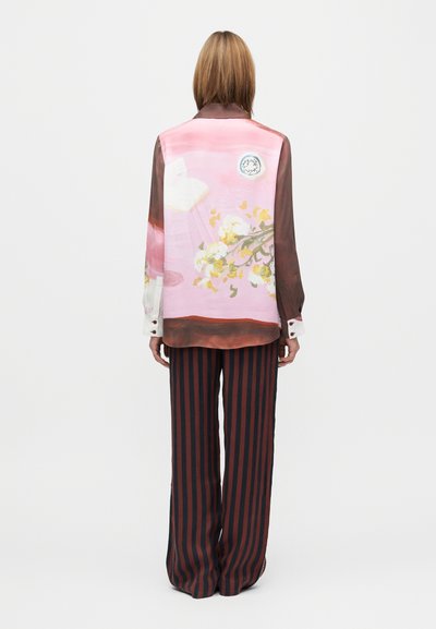 Lengva, permatoma rožinė palaidinė su gėlių raštu ir apvali aplikacija, derinama su platėjusiomis juodai raudonų dryžių kelnėmis. Matomos užsegamos rankogaliai.