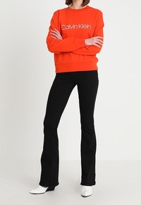 Oranžová mikina s logem "Calvin Klein", doplněná o černé rozšířené džíny a bílé kotníkové boty. Hladký materiál, volný střih a žebrované detaily.