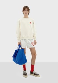 Felpa crema con un punto rosso, pantaloni corti bianchi con un design a cuore rosso, borsa a tracolla blu, calzini rossi e scarpe marroni con suola bianca.