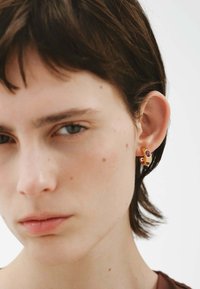 Pendientes en tono dorado con un diseño suave y curvado, y un acento colorido, que muestran un acabado brillante y formas distintivas.