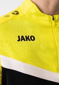 Giacca atletica gialla con un motivo testurizzato. Presenta un logo "JAKO" nero, una striscia bianca e un colletto con zip.