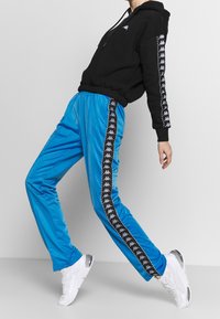 Blå träningsbyxor med elastisk midja, svarta sidostripes med ett logomönster, kombinerade med en svart hoodiesweatshirt. Vita sneakers.