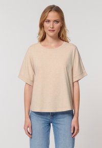 Modella che indossa una t-shirt oversized color pesca chiaro abbinata a jeans blu, in piedi contro uno sfondo neutro, guardando direttamente l'osservatore.