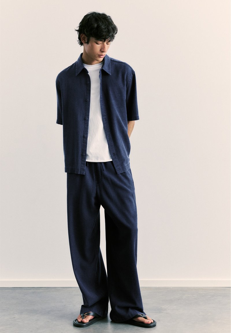 Homme portant une chemise ample bleu marine à manches courtes, un t-shirt blanc, un pantalon large bleu marine et des sandales noires, debout contre un mur beige uni.