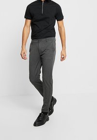 Zwarte rits polo en grijze slim-fit broek, gemaakt van een gestructureerde stof met een knoopsluiting aan de voorkant, gecombineerd met zwarte nette schoenen.