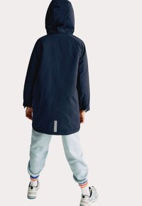 Námorně modrá nepromokavá bunda s kapucí, dlouhá, s nastavitelnými manžetami a reflexním pruhem na zadní straně, v kombinaci s světle modrými tepláky.