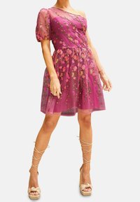 Violetti kukkamekko, jossa on läpikuultavat pönkkähihat, kirjailtuja yksityiskohtia ja istuva yläosa. Päällä beiget korolliset kengät, joissa on ristisidokset.