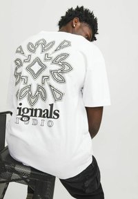 T-shirt bianco con un design geometrico nero sul retro, con le scritte "iginals" e "STUDIO" stampate in grassetto. Vestibilità rilassata, maniche corte.