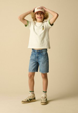 Enfant souriant portant un t-shirt blanc, un short en jean, des baskets et une casquette beige, debout avec les mains sur la tête sur un fond uni.
