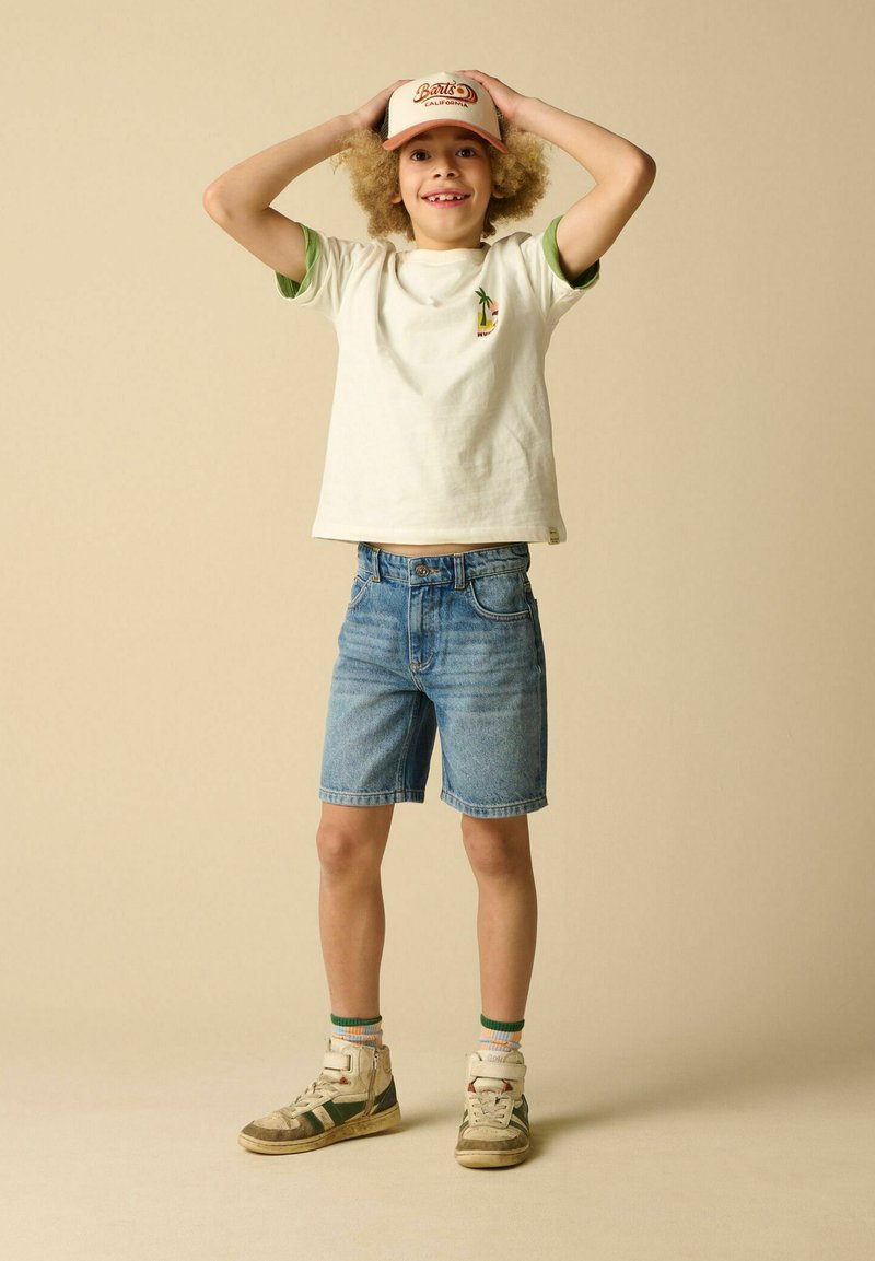 Enfant souriant portant un t-shirt blanc, un short en jean, des baskets et une casquette beige, debout avec les mains sur la tête sur un fond uni.