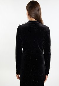 Rochie din catifea neagră cu mâneci lungi, având o suprafață texturată cu detalii reflexive de lumină presărate și butoane decorative pe umăr.