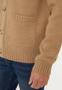 Kamelhaarfarbener Cardigan aus strukturiertem Strickmaterial, mit einer frontalen Tasche, gerippten Bündchen und großen Knöpfen.