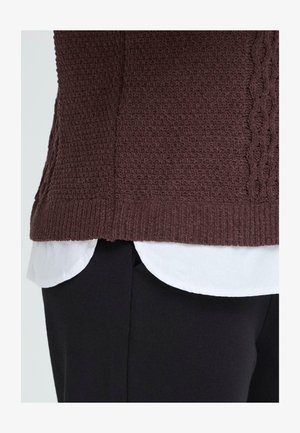 Barna kötött pulóver texturált mintával, fehér ing fölött viselve. Részletei között szerepel bordázott alsó szegély és oldalsó hasítékok. Fekete nadrággal viselve.