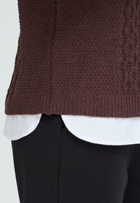 Rjav pleteni pulover z teksturiranim vzorcem, nameščen na beli srajci. Ima rebrast rob in stranske razpore. Nosen s črnimi hlačami.