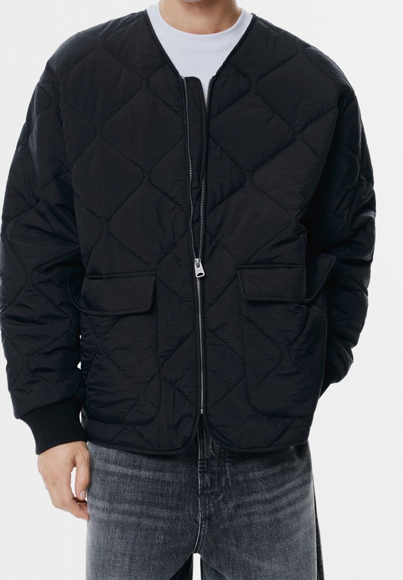 Homme portant une veste matelassée noire avec fermeture éclair devant et poches à rabat, associée à un jean gris et une chemise blanche visible au col.