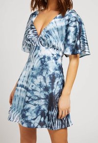 Robe tie-dye bleue avec un col en V profond, des manches flottantes et un corsage ajusté, présentant divers tons de bleu et des motifs abstraits.