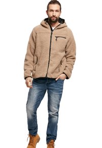 Chaqueta de forro polar color beige con capucha, cremallera negra y dos bolsillos laterales. Lleva puesta una camisa blanca, combinada con unos vaqueros azules y botas marrones.