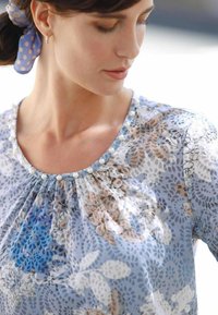 Hellesblaues Blumenshirt mit Rundhalsausschnitt, ausgestattet mit Falten und dekorativen Perlenakzenten entlang des Kragens. Durchscheinender Stoff mit weicher Textur.