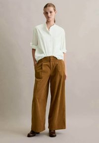 Camisa branca, solta, de botão, com mangas enroladas; calças largas de veludo cotelê castanhas com pregas frontais; botas de cano curto castanhas.