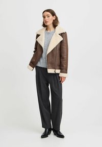 Veste en cuir marron avec col et poignets en shearling crème, associée à un pull gris et un pantalon à carreaux gris. Des bottes noires à la cheville complètent le look.