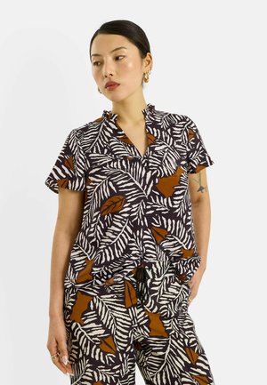 Femme aux cheveux courts et foncés portant une chemise à manches courtes et un pantalon assortis avec un motif noir, blanc et marron, debout avec une main dans la poche.