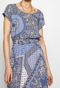 Femme portant une robe bleue à motifs avec une taille smockée et des colliers en or superposés, se tenant devant un fond uni.