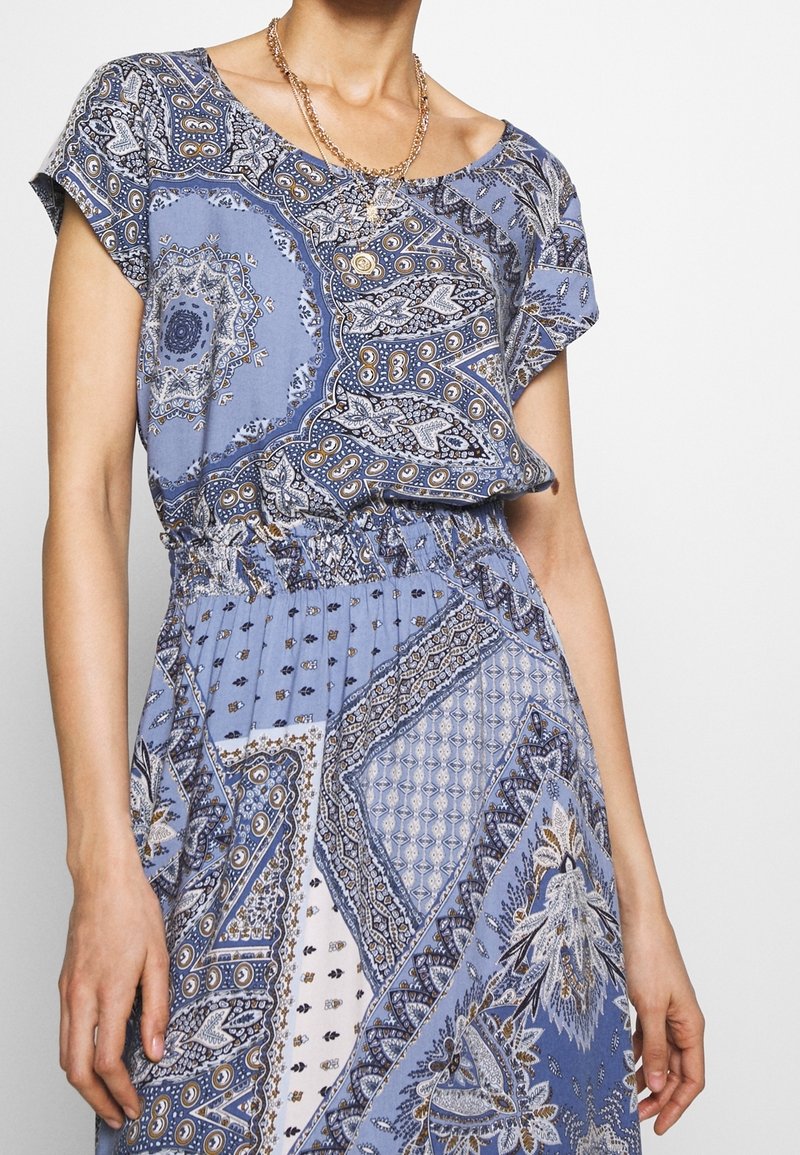 Femme portant une robe bleue à motifs avec une taille smockée et des colliers en or superposés, se tenant devant un fond uni.