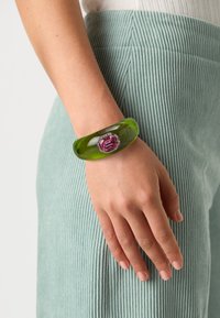 Green acrylic bangle bracelet with a large pink gemstone centerpiece and silver accents, worn on a hand against a textured light green fabric.