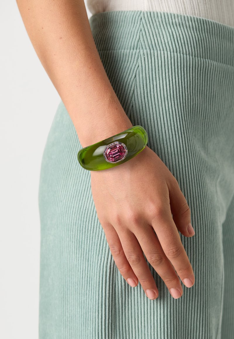 Green acrylic bangle bracelet with a large pink gemstone centerpiece and silver accents, worn on a hand against a textured light green fabric.