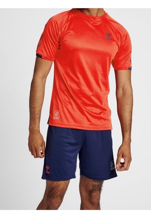 Mann trägt leuchtend orangefarbenes kurzärmeliges Sportshirt und dunkelblaue Shorts, steht vor einem schlichten hellen Hintergrund.