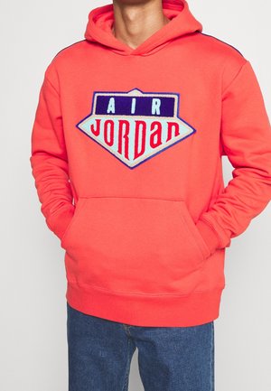 Person iført en koralfarvet Air Jordan hættetrøje med en stor forlomme og blå jeans, hænderne inde i lommen.