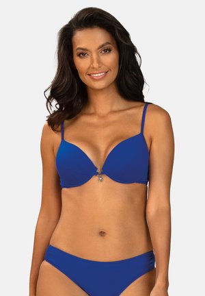 Kobieta z długimi ciemnymi włosami, uśmiechająca się, ubrana w bikini kobaltowe z cienkimi ramiączkami, stojąca na tle jednolitego jasnego tła.