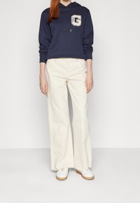 Felpa blu marina con logo, pantaloni larghi crema a vita alta e sneakers bianche. La felpa presenta un cappuccio con cordino e maniche a costine; i tessuti sembrano morbidi.